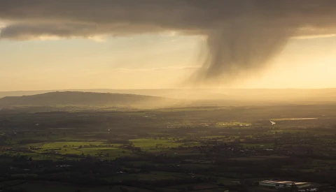 Aerial landscape with rolling hills, patchwork fields, and dramatic storm clouds illuminated by golden sunlight.