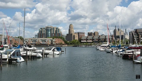 Waterfront marina with sailboats moored alongside a calm harbor, with downtown cityscape and modern buildings in the background.