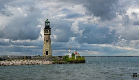 Stone lighthouse with green cupola on rocky breakwater extending into calm waters under dramatic cloudy sky.