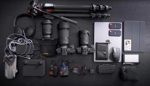 Flat lay of professional video and photography gear including cameras, lenses, audio equipment, and accessories arranged on dark surface.