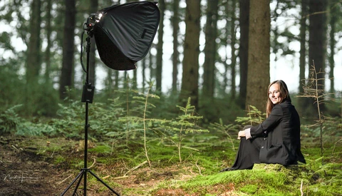 Woman in black blazer seated in forest clearing with professional studio light on stand.