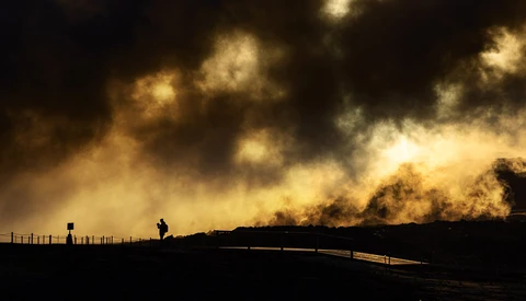 Silhouetted figures standing by a fence with dramatic dust and smoke billowing across a dark sky.