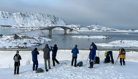 Group of photographers with tripods and equipment standing on snowy ground overlooking a frozen bay with a bridge and snow-covered mountains.