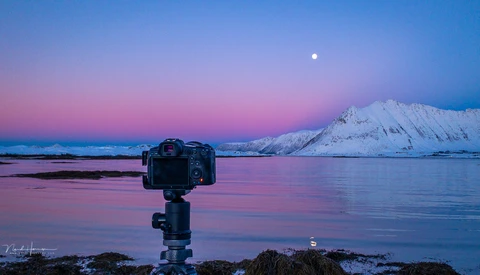 Camera on tripod mounted on rocky shore facing snow-capped mountain across still water at twilight with moon visible.