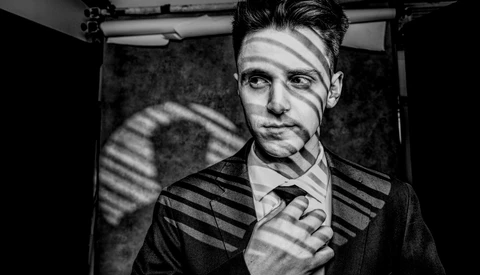 Man in business suit with dramatic window-blind shadows cast across his face and torso.
