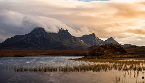 Mountain peaks with low-lying clouds reflected in a still wetland at golden hour.