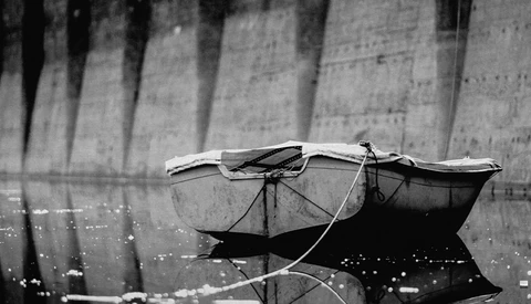 Small fishing boat with furled sail moored against a concrete dam wall, photographed in black and white.