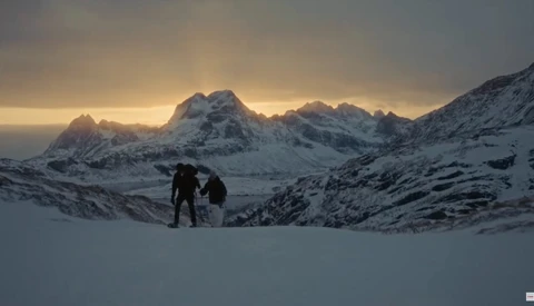 Two hikers stand on snow-covered terrain with snow-capped mountains and golden sunrise light in the background.