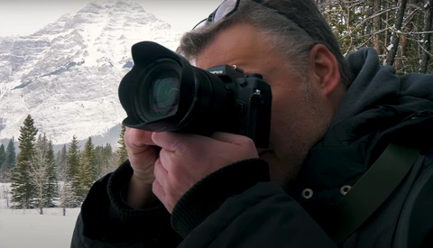 Photographer holding a mirrorless camera with telephoto lens, composing a shot of a snow-capped mountain and forested landscape.