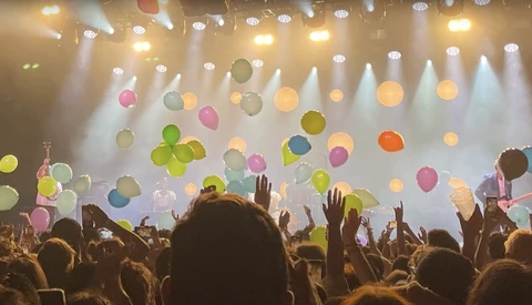 Silhouetted crowd with raised hands at concert with suspended colorful balloons and dramatic stage lighting.