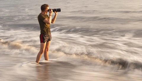 Young photographer wading in ocean waves while holding binoculars to eyes.