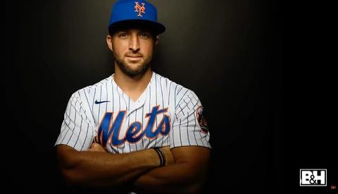 Baseball player in New York Mets pinstriped uniform and blue cap, arms crossed, against dark background.