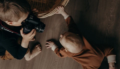 Young child holding a Canon camera while photographing an adult on a wooden floor.