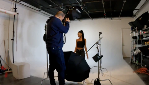 Photographer operating camera on tripod while model in black outfit poses during studio fashion shoot.