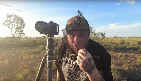 Photographer posing with telephoto lens mounted on tripod in open grassland at golden hour.