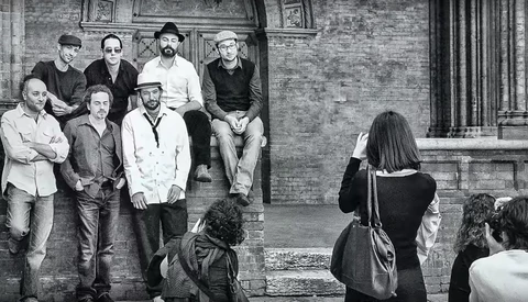 Group of men leaning against brick wall while being photographed by woman in foreground.