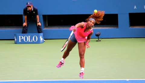 Tennis player in pink outfit executing a serve on a hard court with an umpire seated in the background.