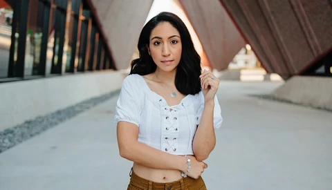 Woman in white lace-up shirt and tan pants holding white hood in architectural hallway.