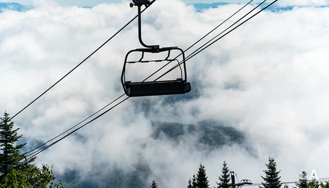 Ski lift chair suspended above clouds with forested mountains partially shrouded in mist below.