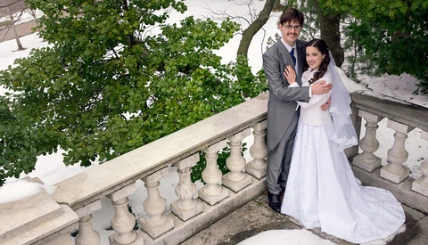 Bride and groom posing together on a stone balustrade with lush green foliage and trees in the background.