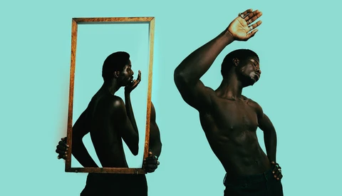 Shirtless man holding wooden frame mirror showing reflection against turquoise background.