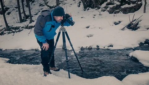 Photographer operating a camera on tripod beside an unfrozen stream in a snow-covered forest.
