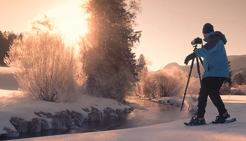 Photographer on skis operating a camera on tripod in snowy landscape at sunrise.
