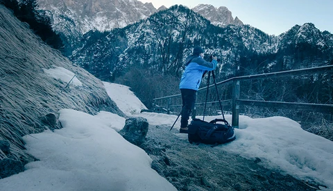 Photographer on a snowy overlook with a tripod and camera, mountains in the distance.