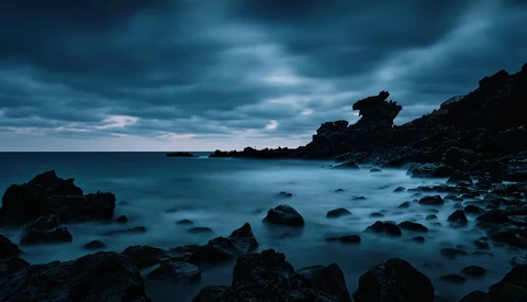 Rocky coastline at twilight with calm, silky water and dramatic storm clouds overhead.