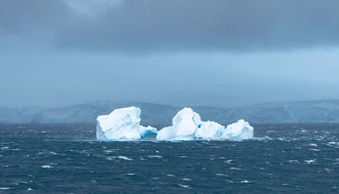 Large blue-white iceberg floating in rough, dark ocean waters with mountains visible in the hazy background.
