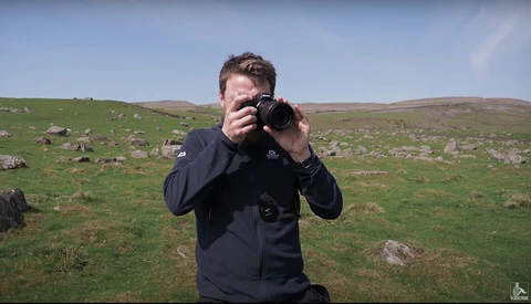 Photographer holding camera to face while standing in open moorland with scattered rocks and green grass.