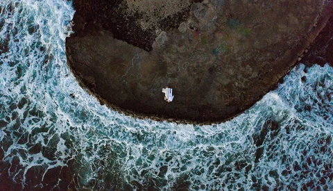 Aerial view of a small white boat in a dark cove surrounded by turquoise ocean waves and rocky coastline.