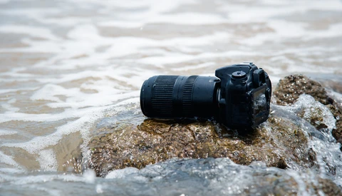 Black DSLR camera with telephoto lens resting on a rocky outcrop in shallow ocean waves.