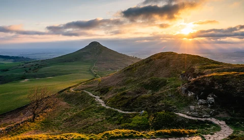Conical mountain with winding path under golden sunset sky, distant valley landscape below.