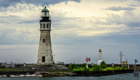 Stone lighthouse with green cupola stands on rocky breakwater under dramatic cloudy sky.