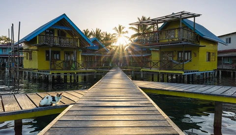 Symmetrical wooden boardwalk extending toward two bright yellow stilt houses with blue roofs flanking palm trees at sunrise.