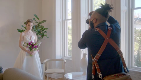 Photographer adjusting camera settings while bride in white dress holds purple bouquet in bright room.