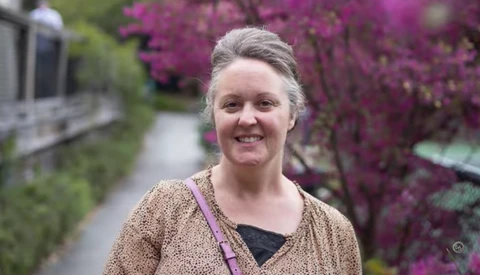 Woman smiling outdoors with vibrant purple flowering shrubs in soft, natural daylight.