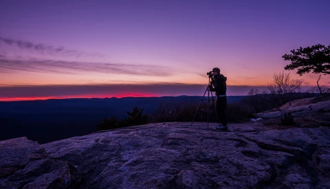 Photographer on rocky outcrop silhouetted against vibrant purple and pink sunset sky.