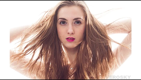 Woman with long brown hair and magenta lipstick, hands raised to head against bright backlit background.