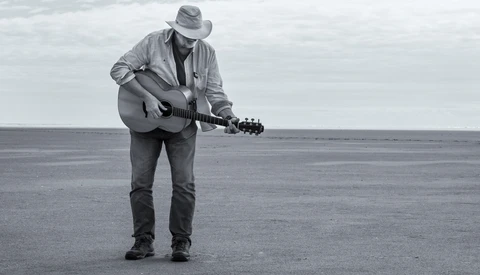 Musician playing acoustic guitar on a vast, empty beach with overcast sky.