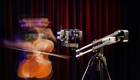 Motion-blurred cellist performing in front of a camera mounted on a tripod against a red curtain backdrop.