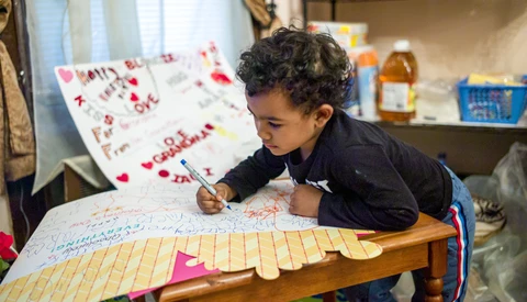 Child writing on paper at a wooden table with a xylophone visible in the foreground.