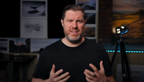 Man in black t-shirt speaking to camera in a studio setting with professional lighting equipment visible.