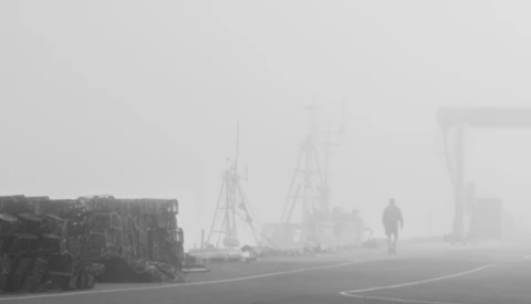Foggy industrial harbor with a silhouetted figure standing on a wet road among cranes and stacked materials.