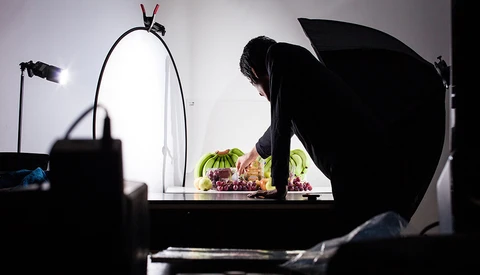 Photographer setting up a still life shot of fresh fruits under studio lighting with reflectors and key light positioned around the setup.