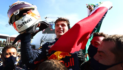 Racing driver celebrates with team members holding a large red flag after a victory.