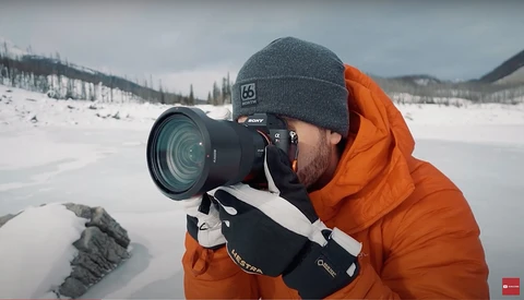 Photographer in orange jacket and gray beanie holding DSLR camera with telephoto lens while shooting on snow-covered landscape.