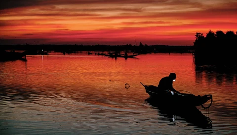 Silhouetted fisherman in small boat on calm water at dramatic sunset with fiery orange and red sky.