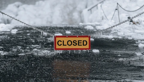 Red and yellow CLOSED sign suspended by chains over an icy river with snow-covered banks.